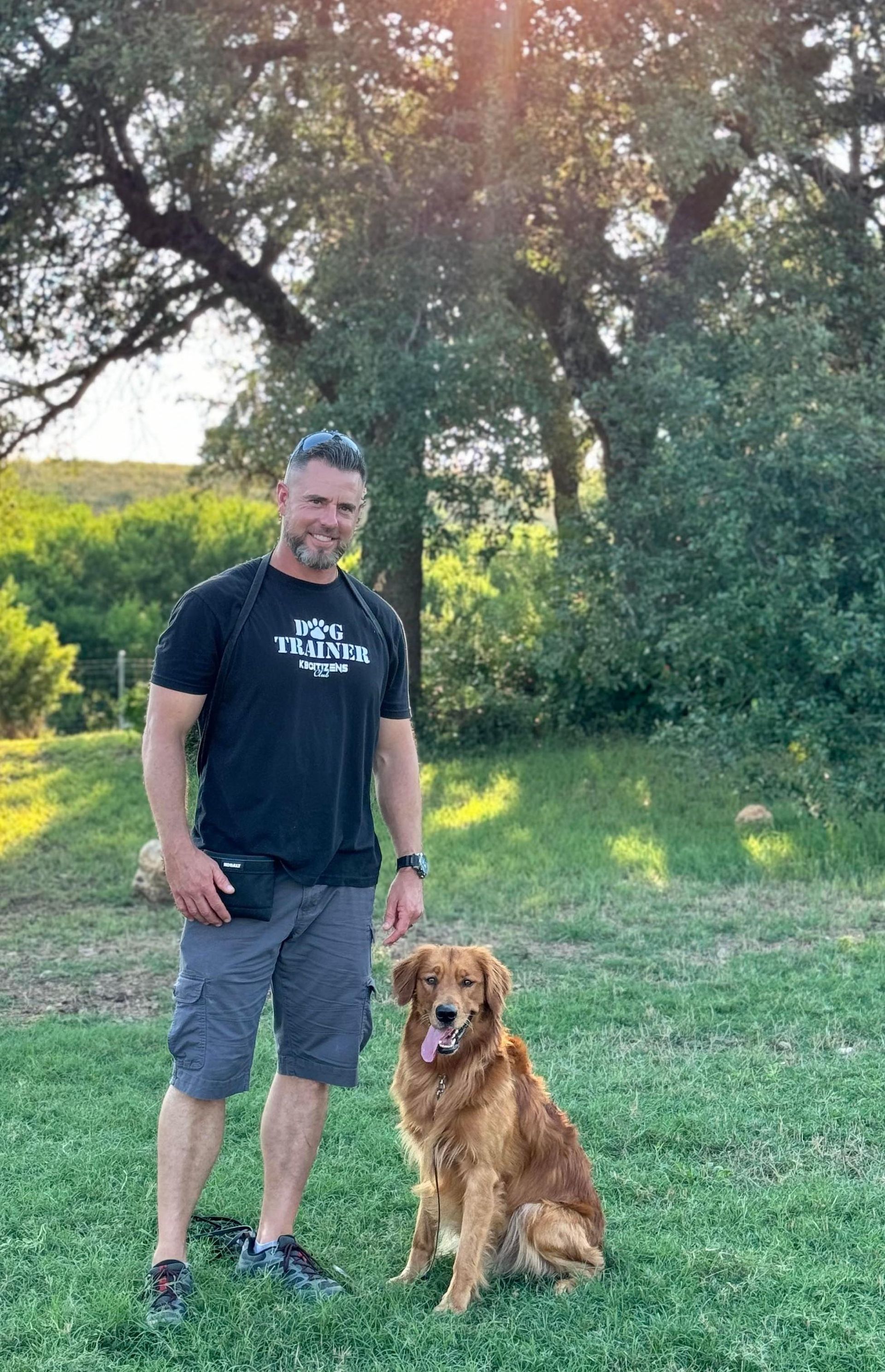 Man in black shirt and gray shorts with golden retriever sitting in a grassy yard, sunlit trees in the background.