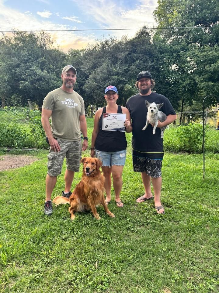 Three people and two dogs pose outdoors. Woman holds paper, golden retriever sits, and small dog is held.