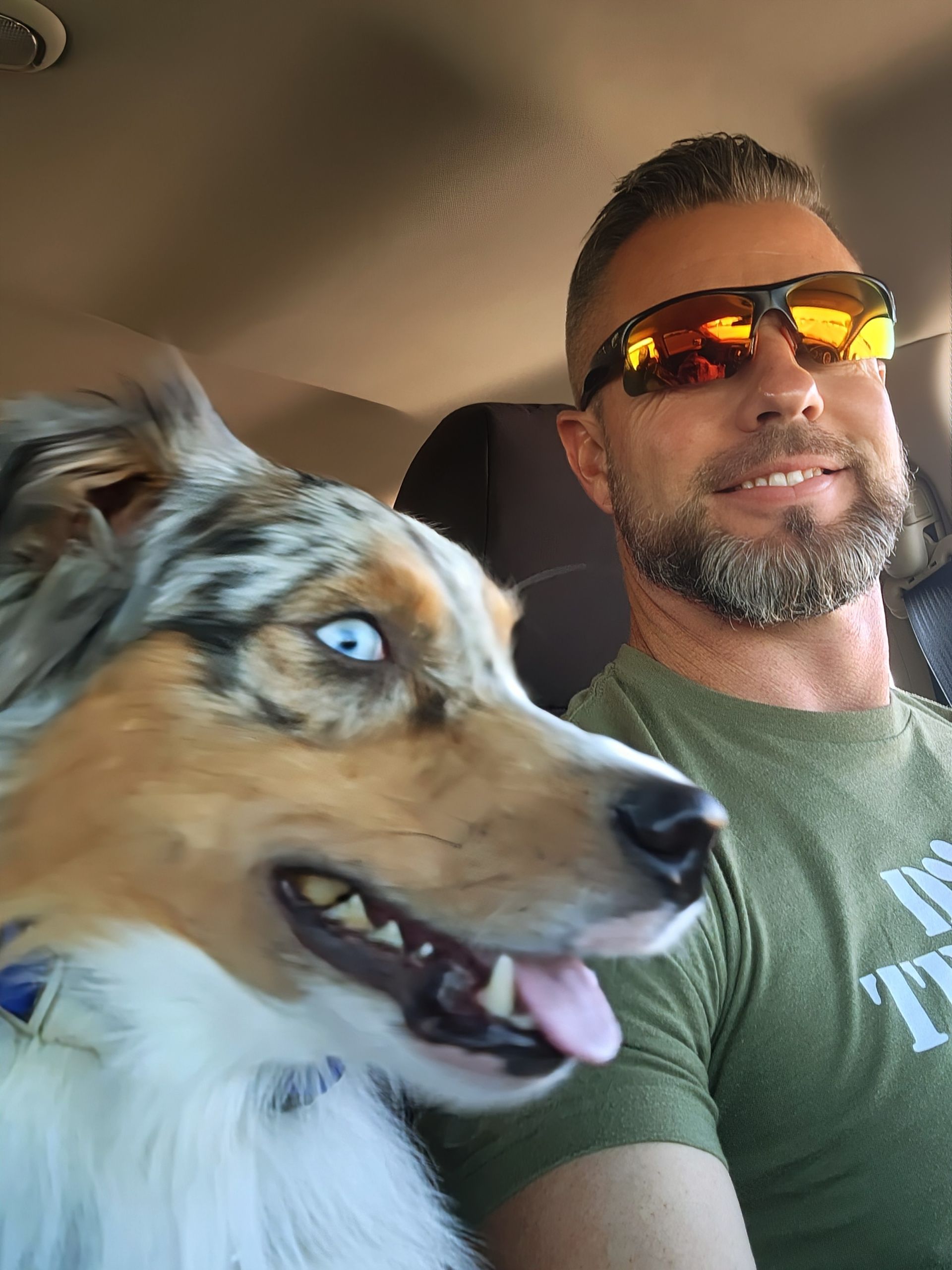 Man wearing sunglasses with dog in car, both looking ahead. Dog's blue eyes visible.