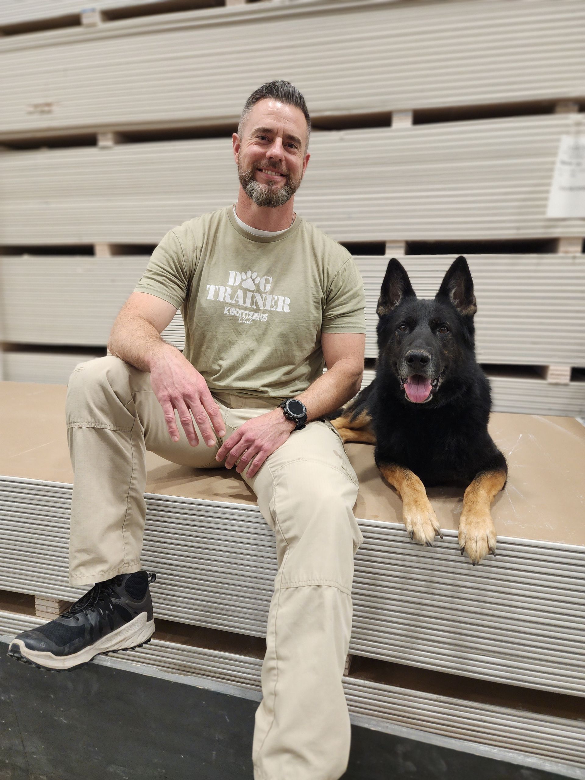 Man in khaki with a black and tan dog sitting in a warehouse.