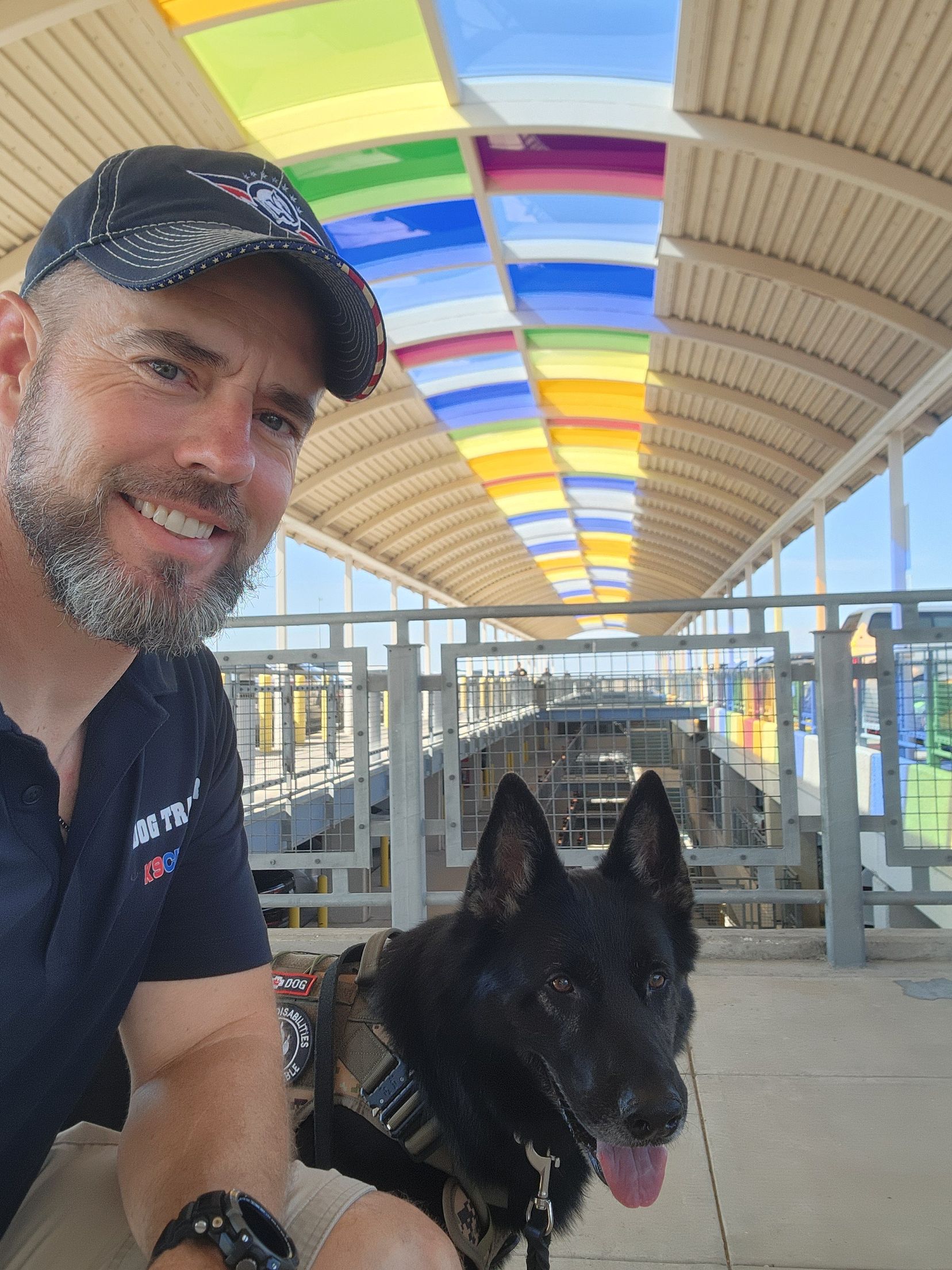 Man with black dog smiles at camera; under colorful, arched walkway.