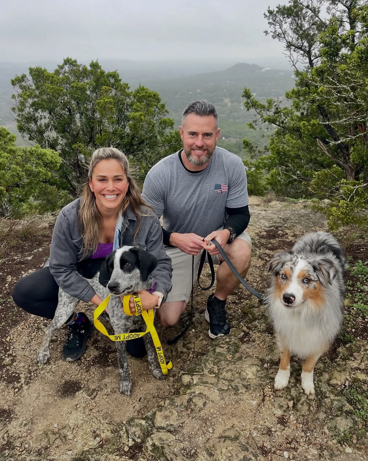 A couple kneels with two dogs on a rocky hilltop with a distant, misty view.