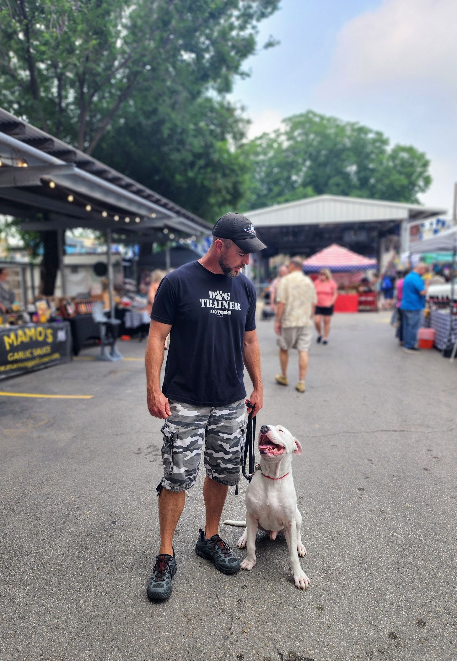 Man in black shirt and camouflage shorts with white dog on leash outdoors. Market setting.