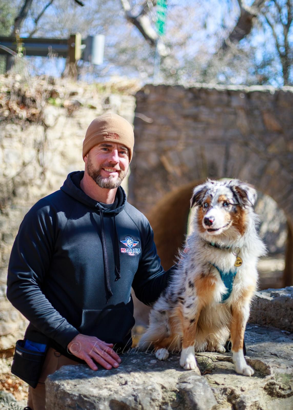 Man in black hoodie and beanie, with an Australian Shepherd dog outdoors in front of stone arch.