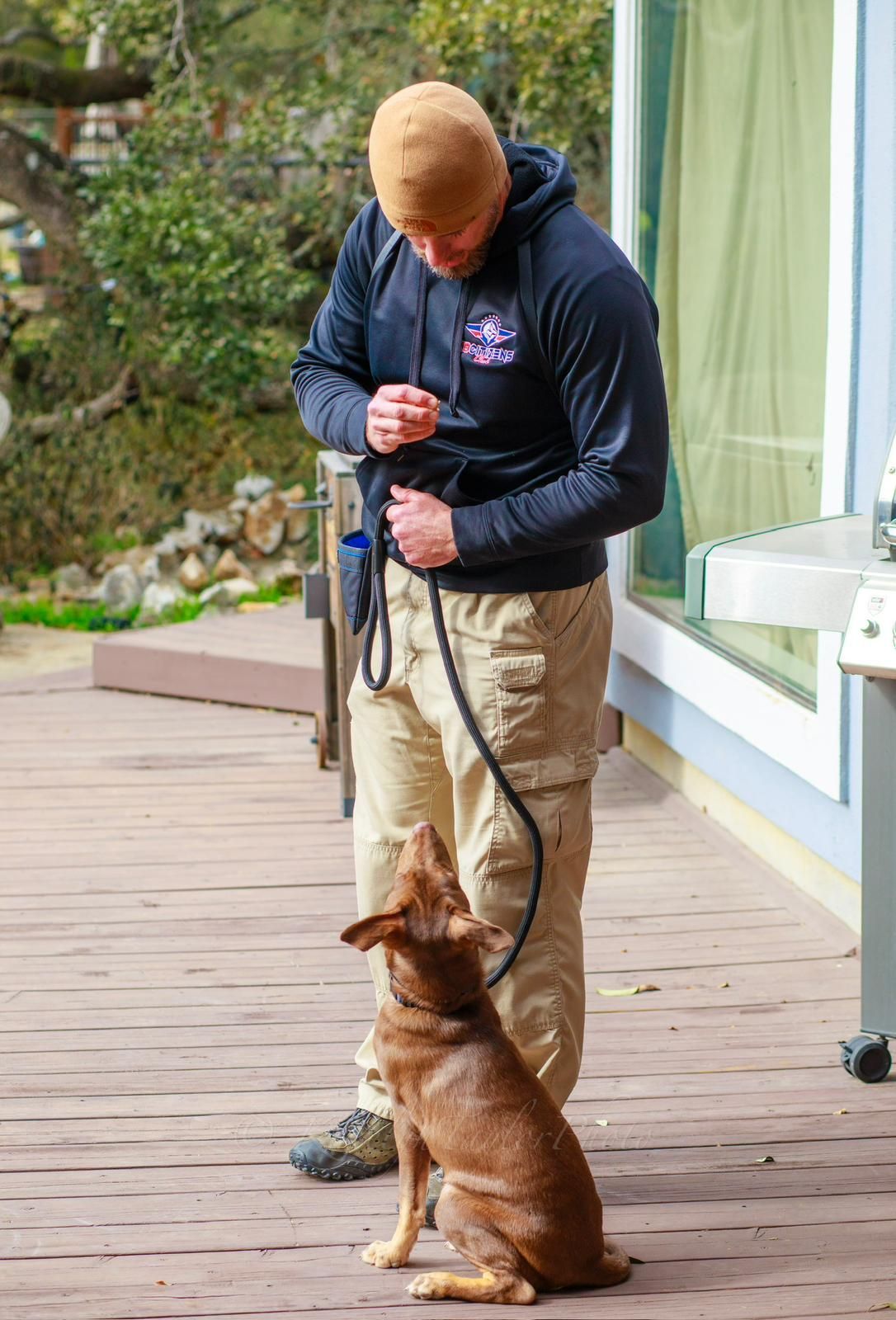 Man in black hoodie and khaki pants training a brown dog on a wooden deck.