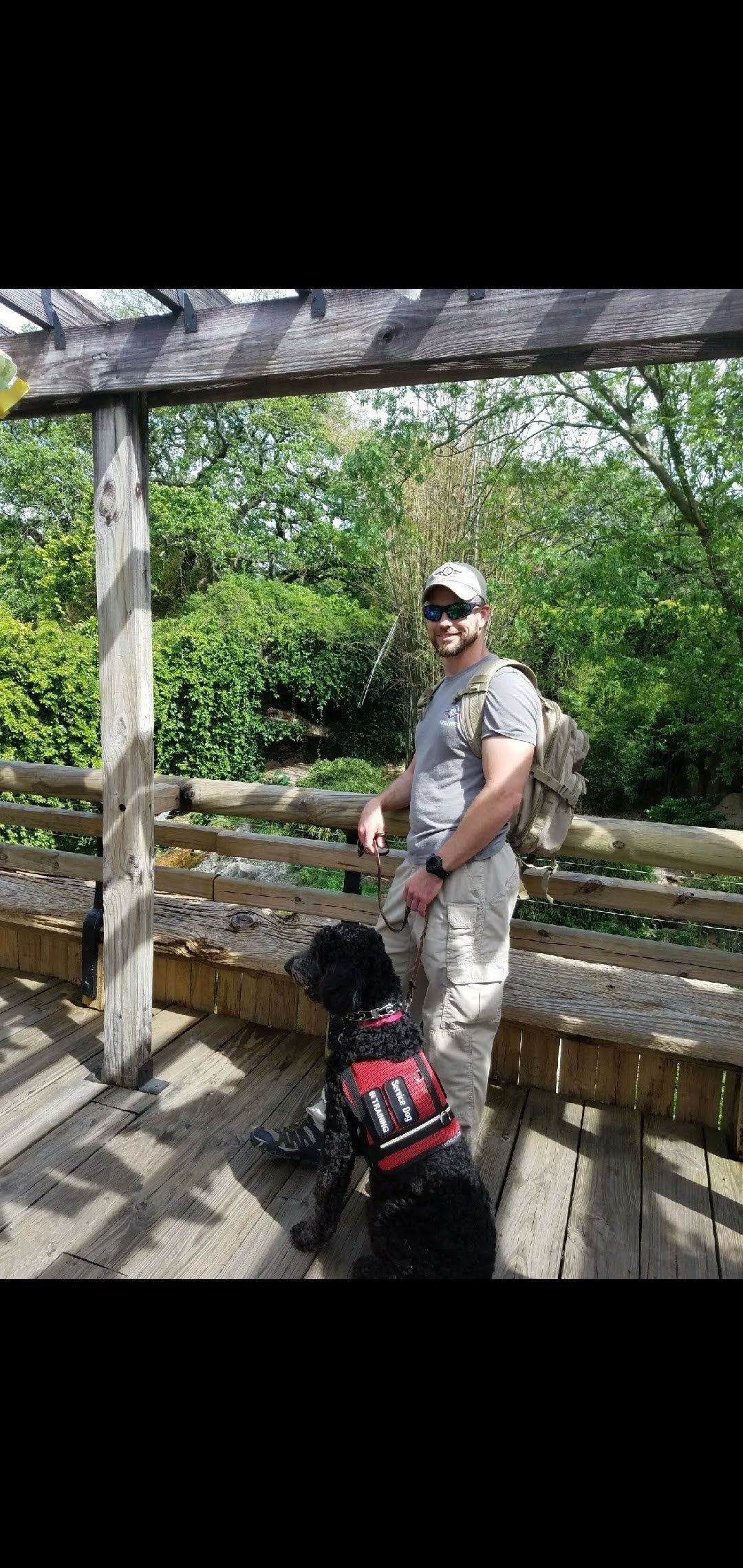 Man with a black dog on a wooden deck. Dog wears a red vest. Trees in background.