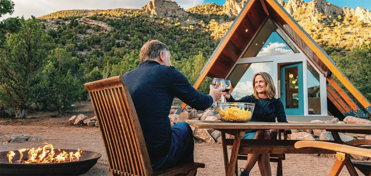 A man and a woman are sitting at a table with wine glasses in front of an a frame cabin.