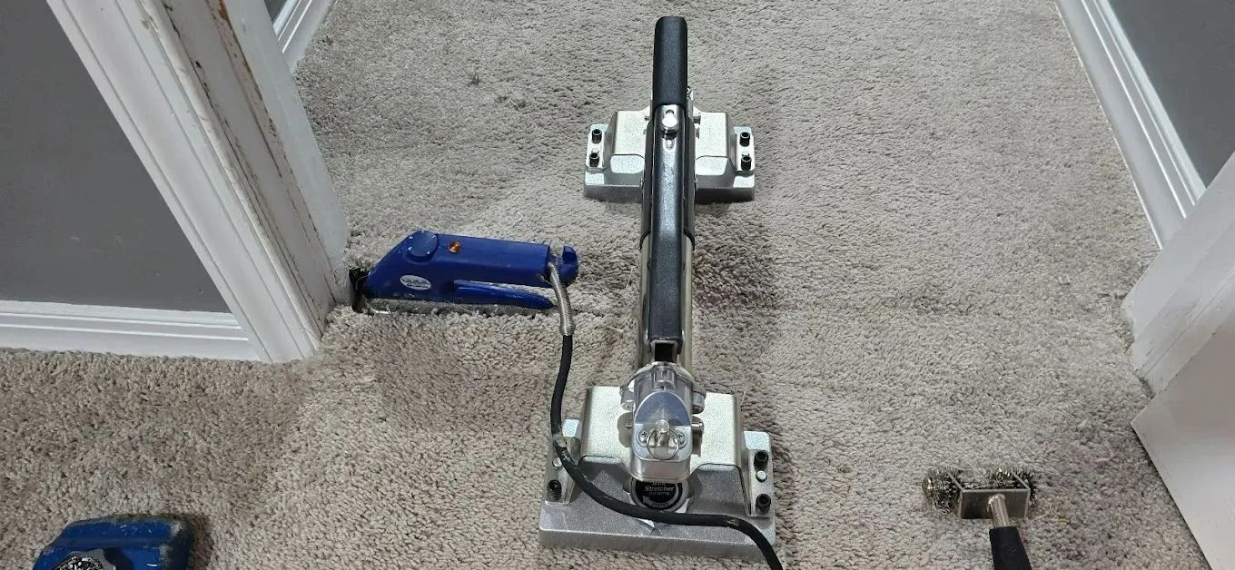 Carpet installation tools, including a seam iron and power stretcher, are positioned on light-colored carpet in a doorway.