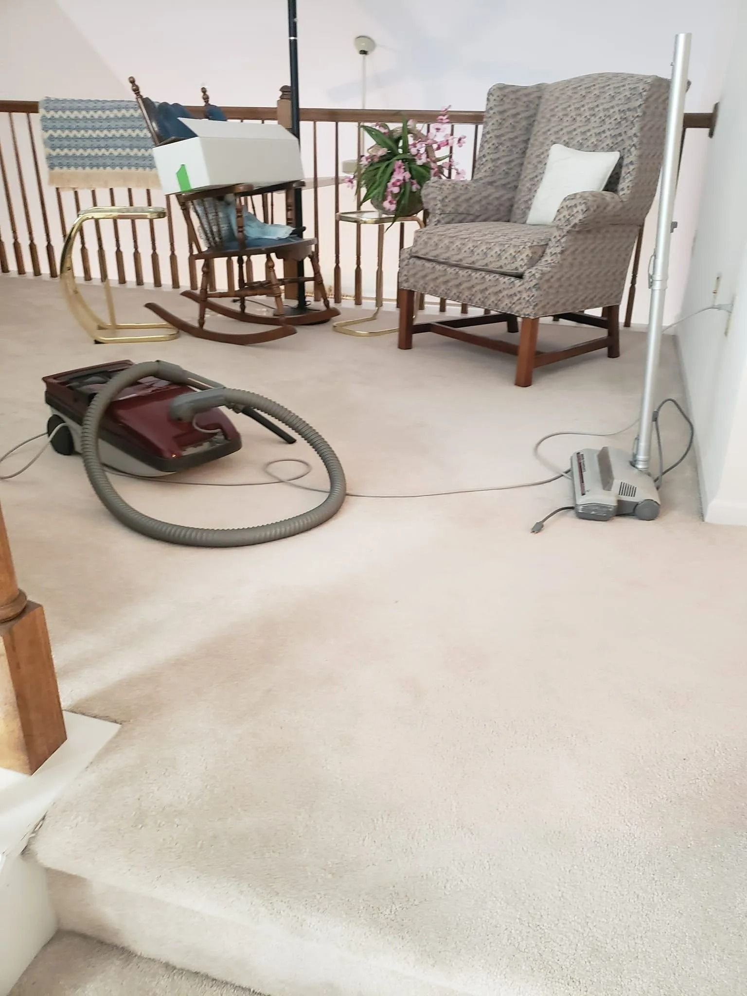 A vacuum cleaner sits on a carpeted landing near a rocking chair and an upholstered armchair by a wooden railing.