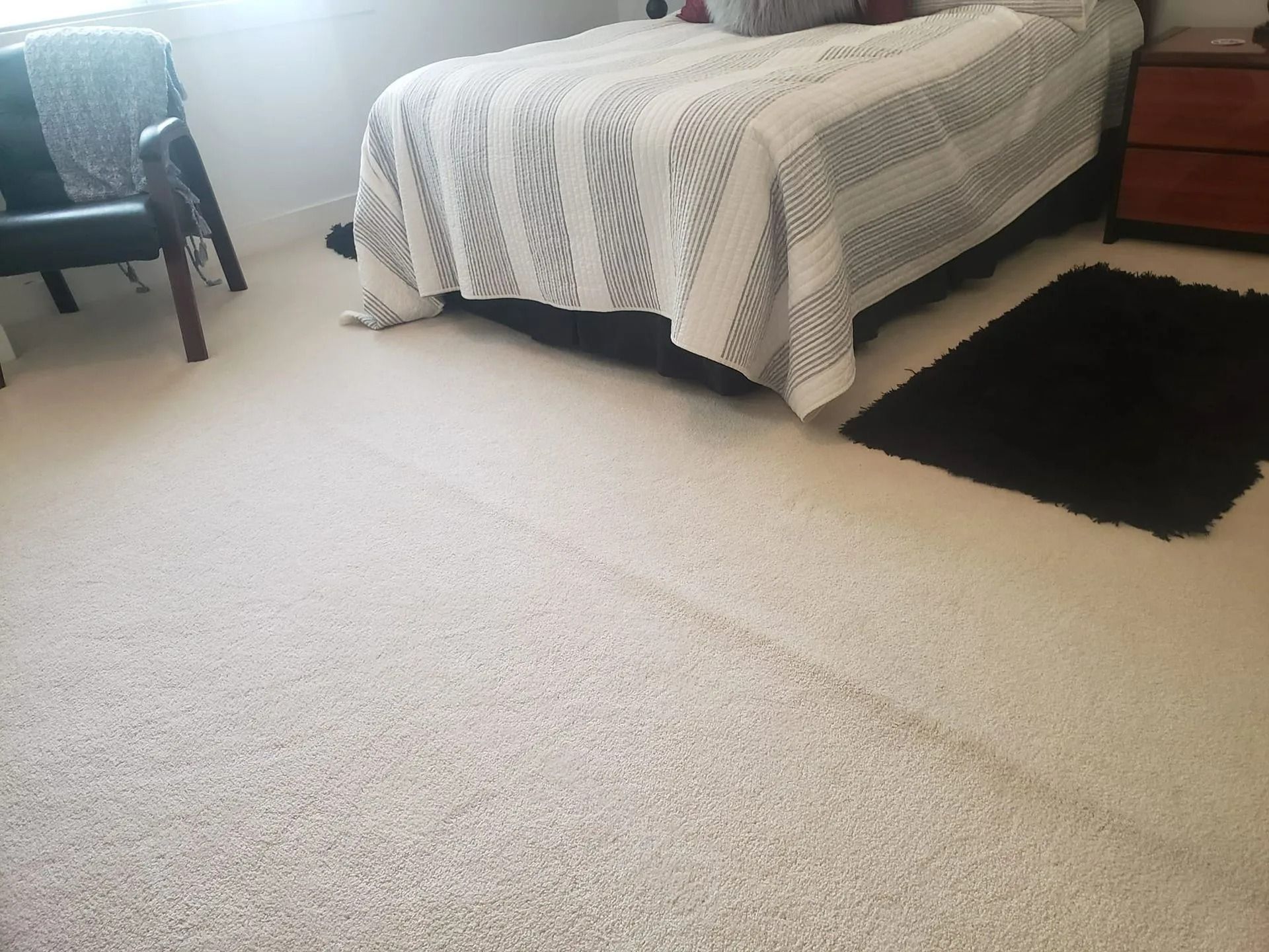 A bedroom featuring light carpeting, a bed with a patterned cover, a dark armchair, and a black rug on the floor.