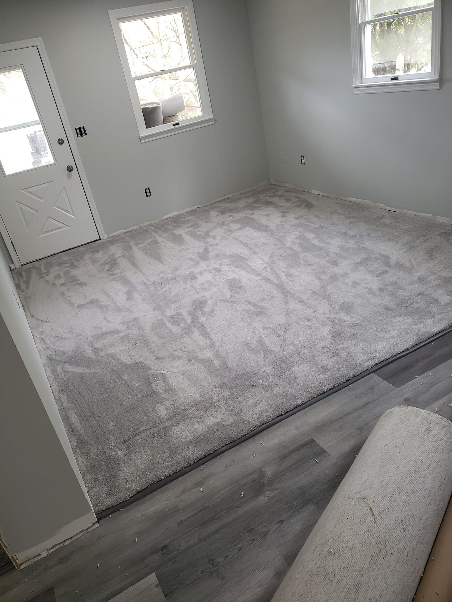 A room transition showing plush light gray carpet meeting gray wood-look flooring, with a white door and two windows.