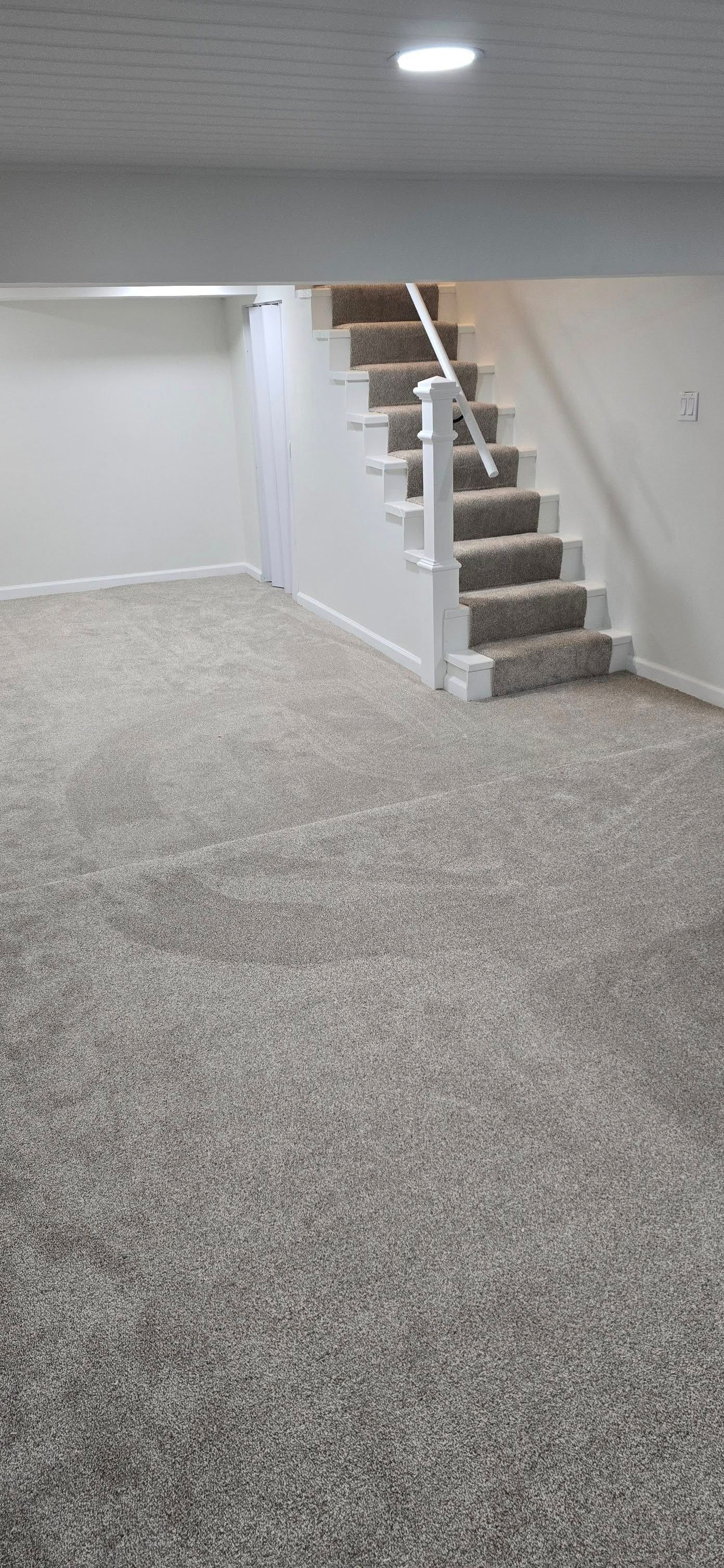 A finished basement with neutral gray carpet and white walls featuring stairs leading to the floor above.