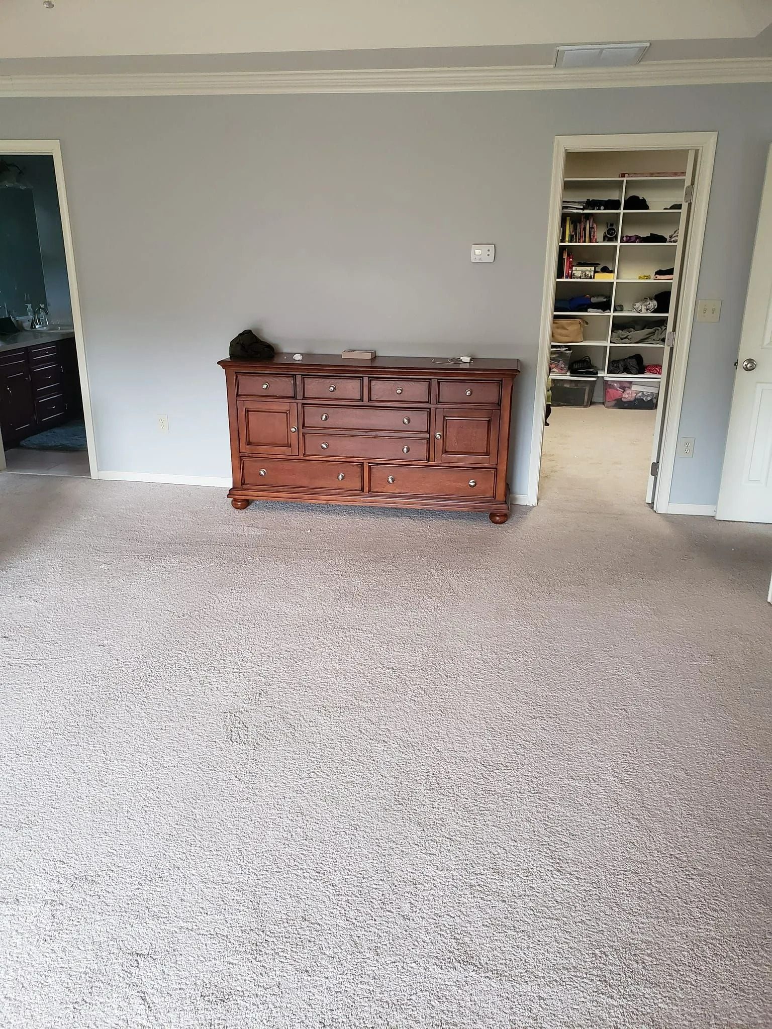 A wooden dresser stands against a gray wall between two doorways, with patterned wall-to-wall carpeting in a room.