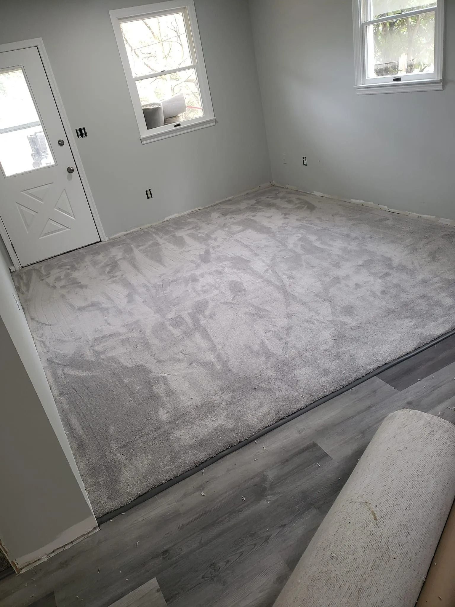 A room with gray carpet installed, transitioning to gray wood-look flooring near the foreground with a carpet roll.