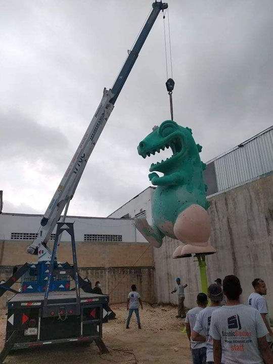 Una grúa levanta una gran estatua de dinosaurio verde, con aspecto de caricatura. Los trabajadores observan la instalación cerca de un edificio.