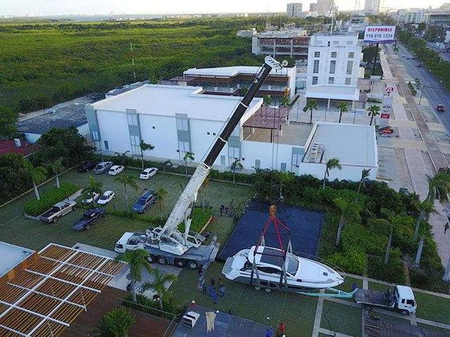 Una grúa levanta un yate desde un remolque en un área de estacionamiento cerca de edificios y follaje.