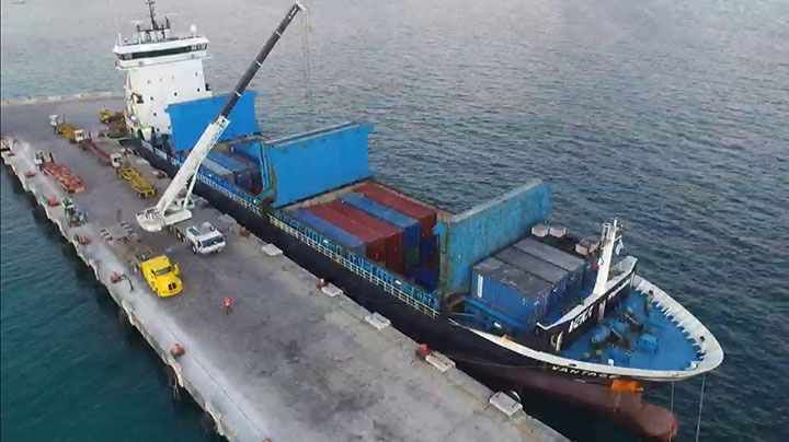 Buque de carga atracado en un muelle, siendo descargado por una grúa; contenedores azules, grúa blanca, agua azul.