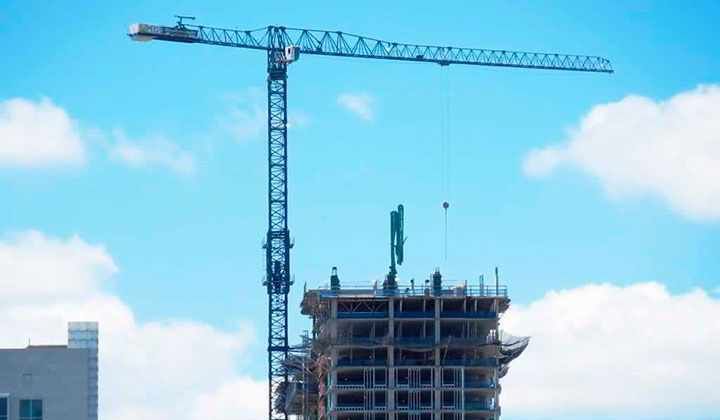 Grúa de construcción azul levanta material a un edificio de varios pisos en construcción contra un cielo azul con nubes.