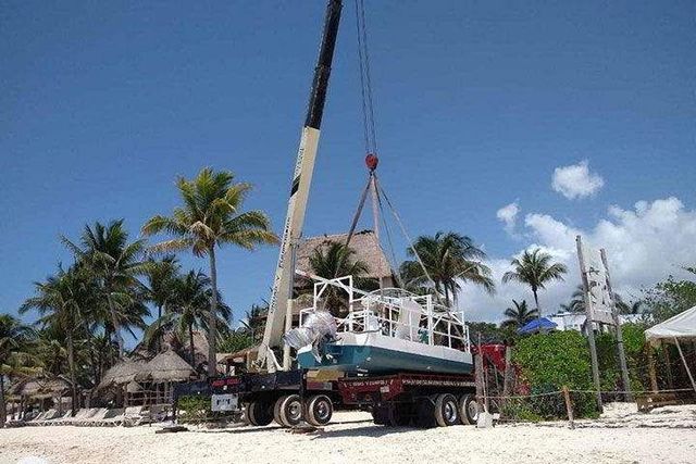 Una grúa levanta un barco desde un camión en una playa con palmeras y un cielo azul claro.