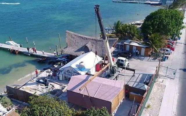 Una grúa eleva un componente de un edificio junto a una playa y un muelle. Un pequeño edificio se encuentra debajo del componente y se ve el mar.