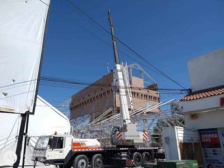 Una grúa levanta una gran estructura metálica reflectante cerca de un edificio. También se ven el cielo azul, cables y un camión blanco.