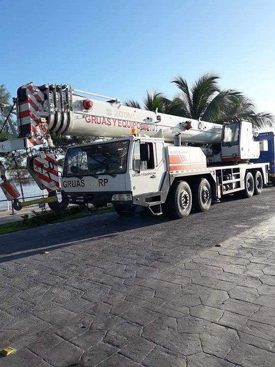 Camión grúa blanco estacionado en un área pavimentada, con la pluma extendida, cerca de agua y palmeras.