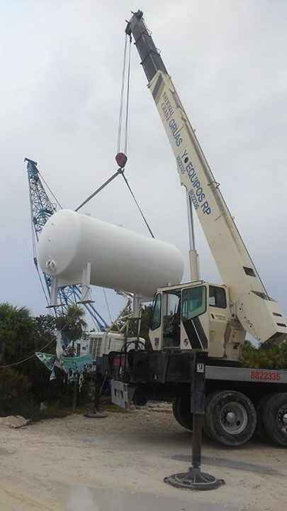 Un gran tanque blanco es levantado por una grúa en un día nublado. Se ve otra grúa al fondo.