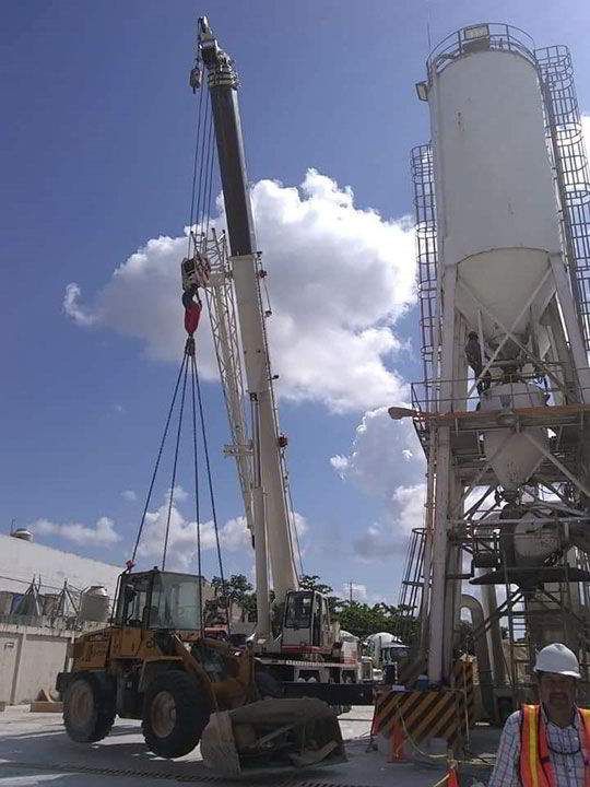 Grúa levantando un cargador frontal amarillo en un sitio de construcción cerca de un silo bajo un cielo parcialmente nublado.