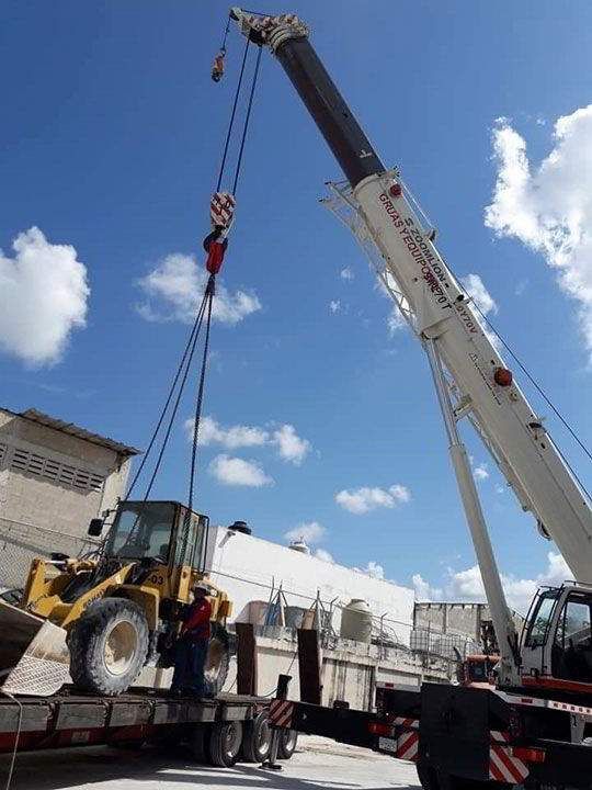 Una cargadora frontal amarilla es levantada por una grúa desde un camión de plataforma en un día soleado.