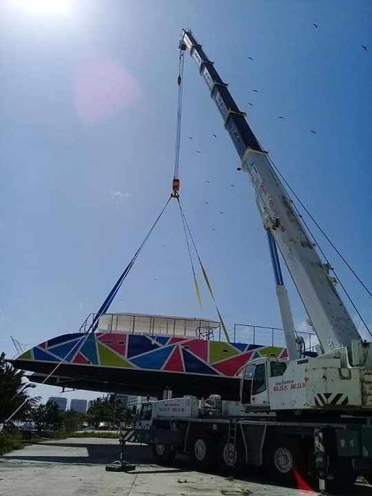 Una grúa levanta un barco colorido contra un cielo azul brillante.