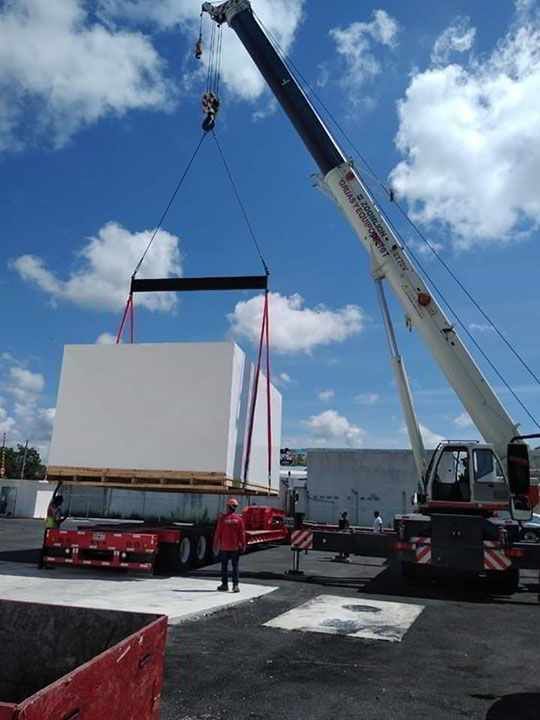 Una grúa levanta una gran caja blanca de un remolque de plataforma en un sitio industrial.