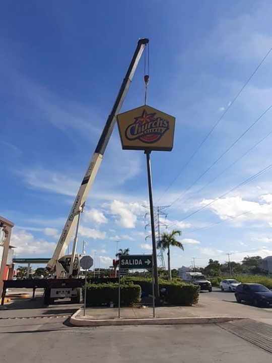 Una grúa levanta un cartel de Charleys Philly Steaks al borde de la carretera. Se ve el cielo azul, cables de electricidad y algunos vehículos.