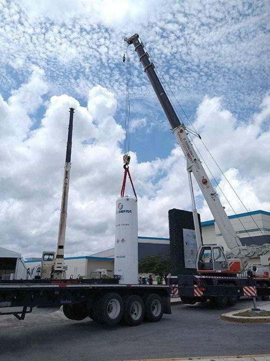 Dos grúas levantan un objeto cilíndrico blanco desde un camión de plataforma contra un cielo nublado.