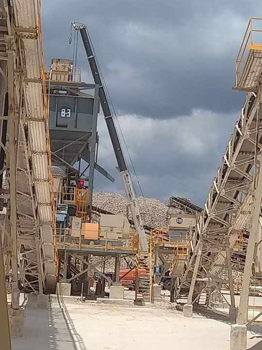 Grúa en una cantera, elevando equipo. Estructuras grises y canela con cintas transportadoras contra un cielo nublado.