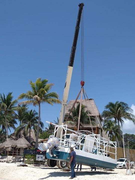 Grúa levantando un barco blanco y azul en una playa, cerca de una estructura de madera y palmeras. Día soleado.