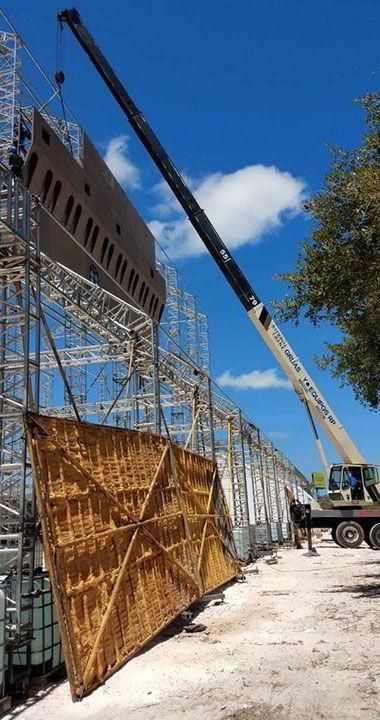 Una grúa levanta un gran panel geométrico junto a un andamio bajo un cielo azul.