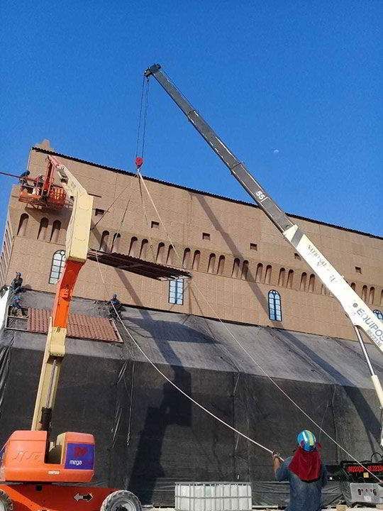 Una grúa levanta un objeto grande cerca de un edificio de ladrillos. Una persona guía cuerdas y otra en un ascensor. Cielo soleado.