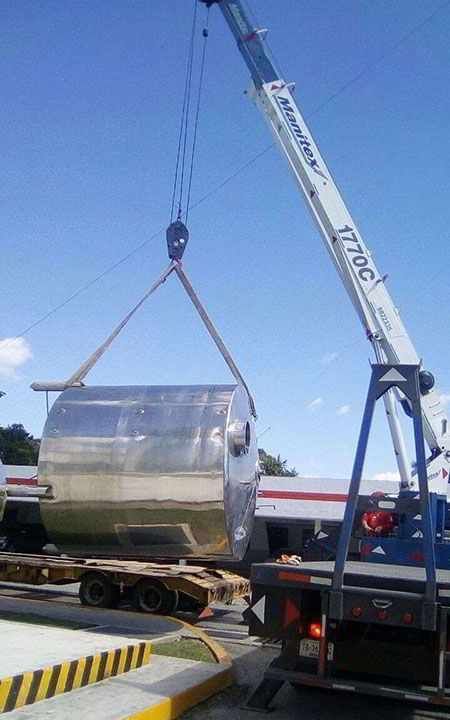 Grúa levantando un gran objeto cilíndrico plateado desde un remolque de plataforma en un día soleado.