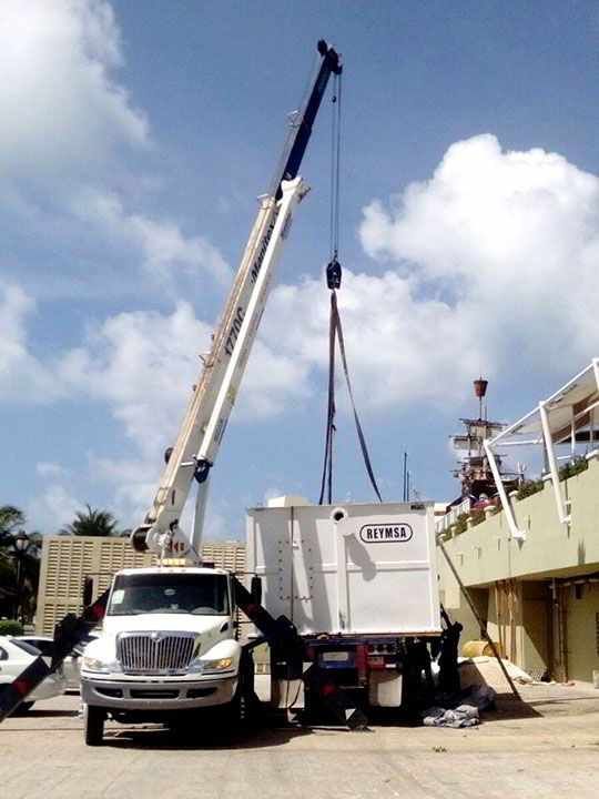 Grúa blanca levantando un gran contenedor blanco desde un camión de plataforma cerca de un edificio.