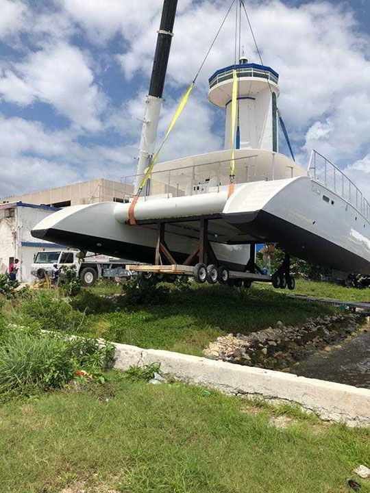 Un catamarán está siendo levantado por una grúa cerca de un edificio con una torre, probablemente en un puerto deportivo.