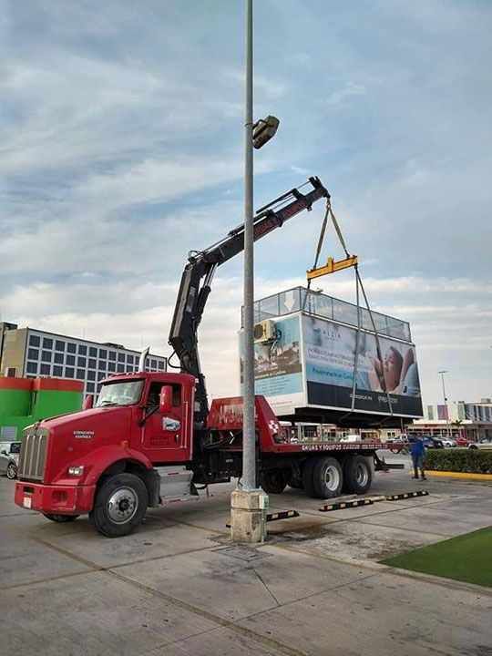 Grúa de camión rojo levantando una gran exhibición rectangular con una escena de playa, estacionada sobre el pavimento, cielo nublado.