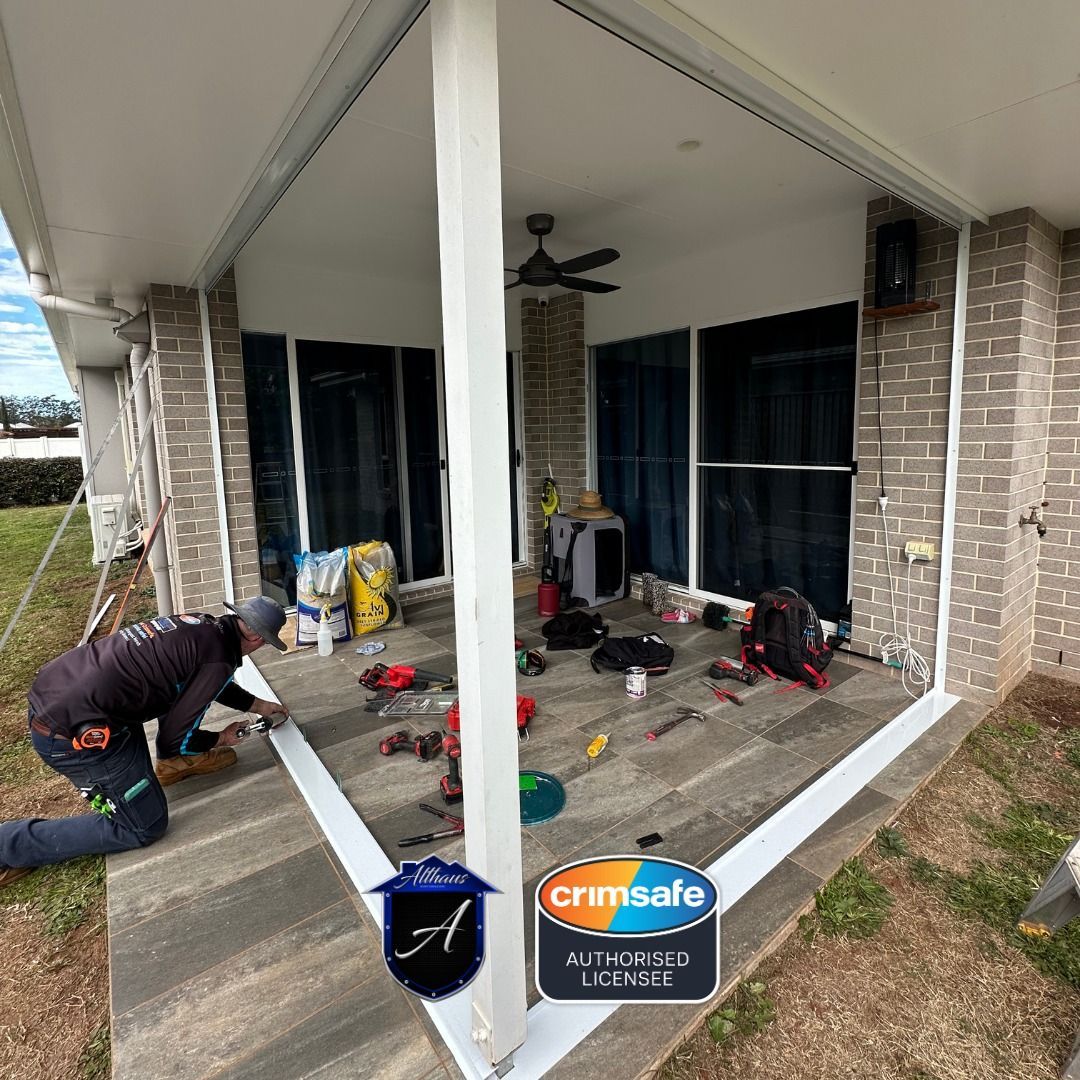 A worker kneeling on a tiled patio, installing white framing for a Crimsafe security screen system at a house.