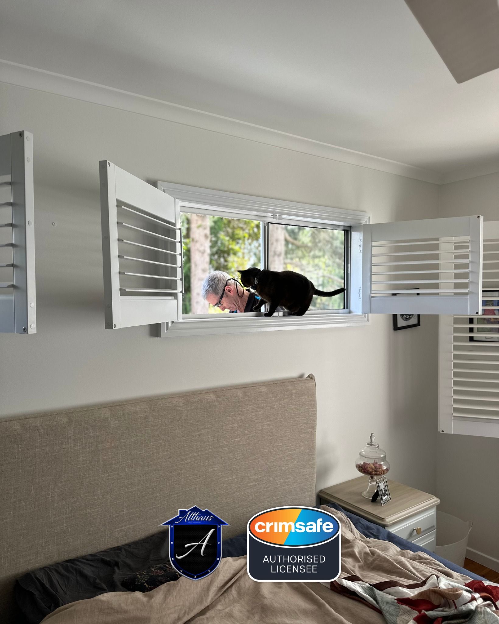 A person peeks through an open window at a black cat sitting on the sill in a bedroom.