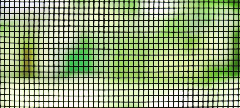 A close-up view of a dark window screen mesh with a blurry, bright green and white outdoor background. A close-up view of a dark window screen mesh with a blurry, bright green and white outdoor background.