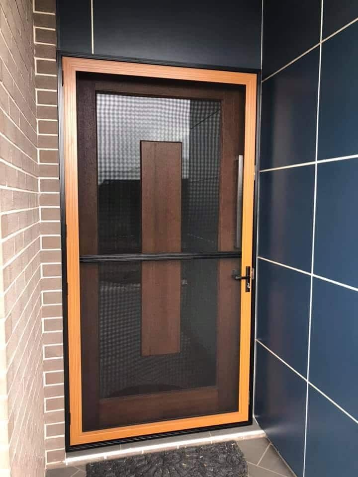 A modern screen door with a wooden frame and black mesh, set in an entrance between a brick wall and dark blue panels.