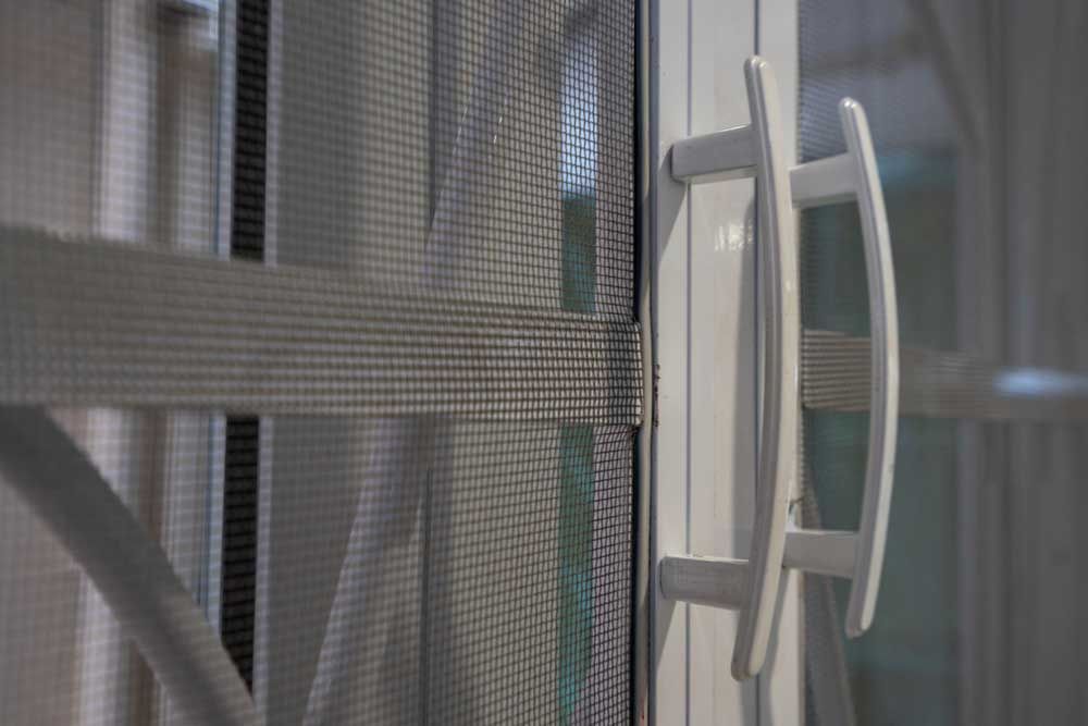 A close-up view of a white door handle attached to a white sliding door frame with a metal mesh screen.