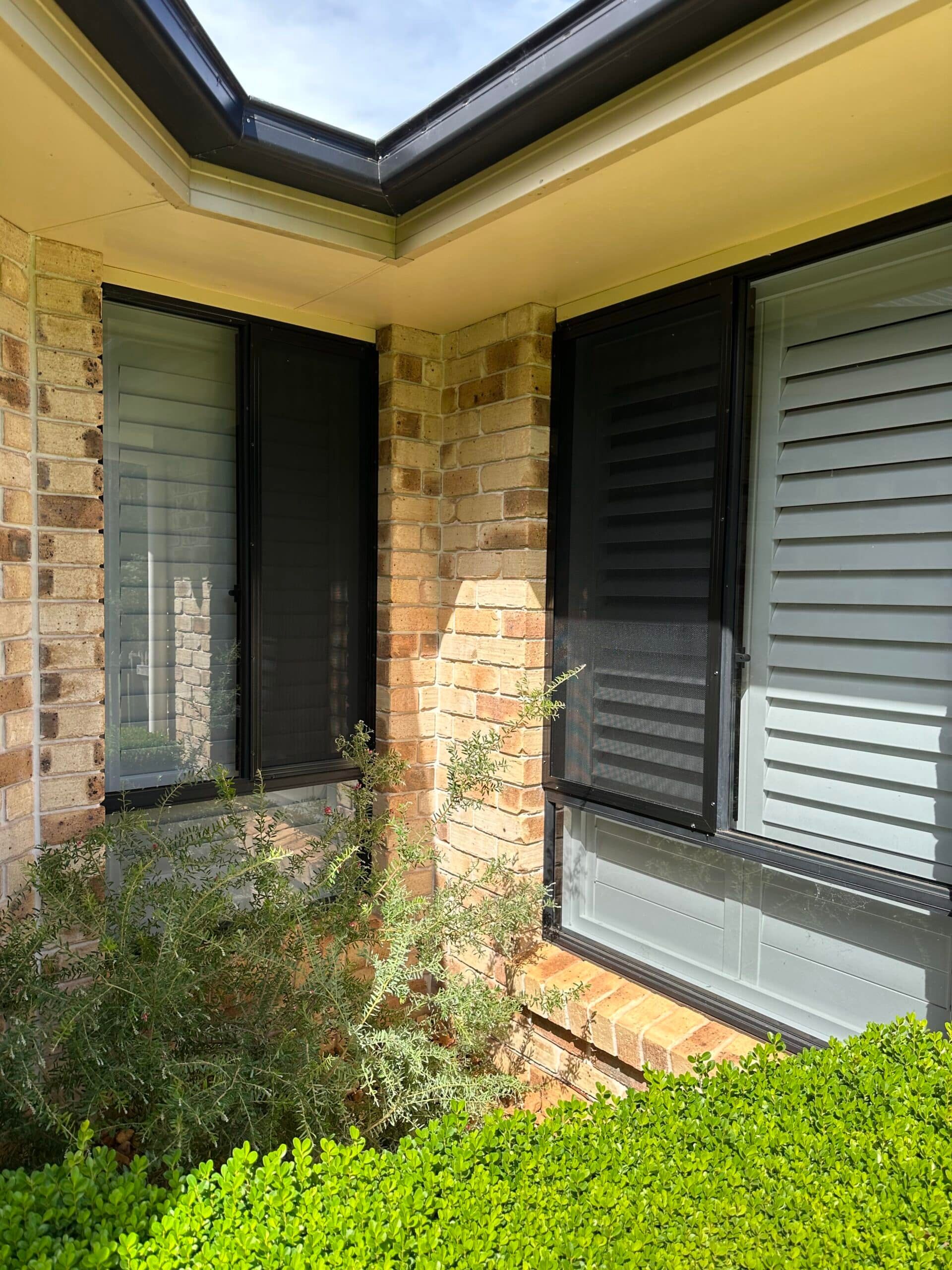 Corner view of a brick house exterior with dark-framed windows, security screens, and green garden hedges below.