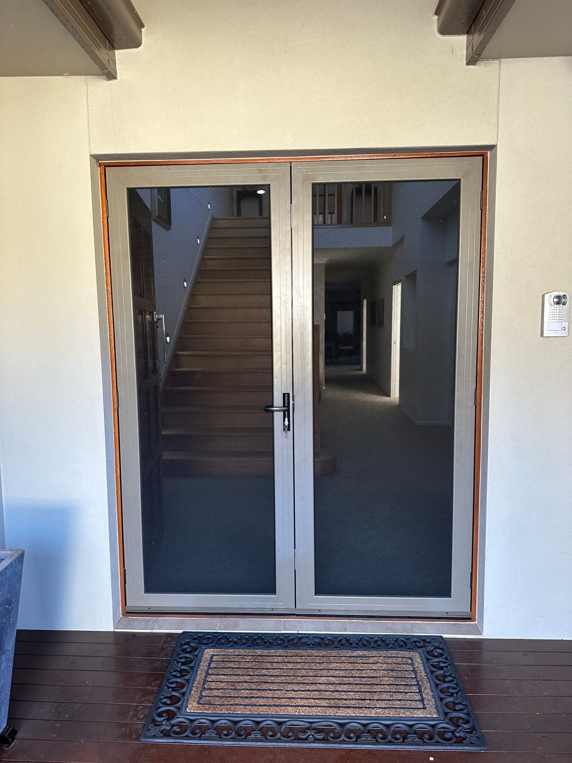 A neutral-colored double screen door on a porch, framed by a beige wall with a doormat in the foreground.