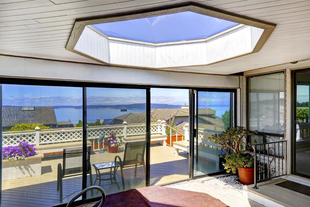 A bright sunroom with a hexagonal skylight overlooks a patio with a view of the ocean and distant hills.