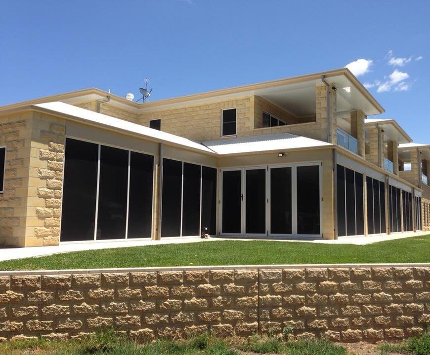 A two-story house with light tan stone walls, large glass windows with dark screens, and a terraced stone retaining wall.